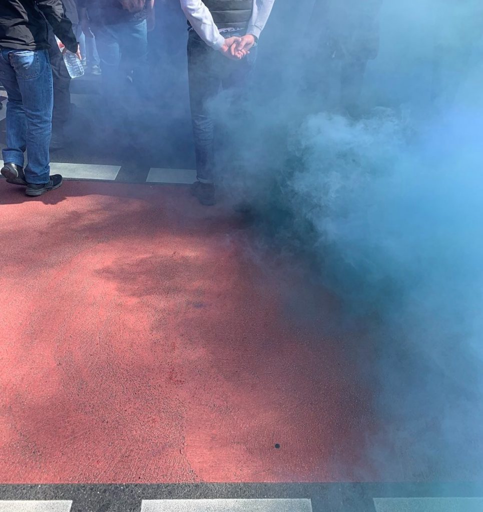 Straßenboden, eingenebelt von blauem Rauch, am oberen Bildrand sieht man Menschenstehen