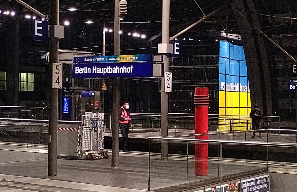 Blick auf ein Bahnhofsgleis. "Berlin Hauptbahnhof". Im Hintergrund ein blau-gelbes Banner mit der Aufschrift "#StandWithUkraine"
