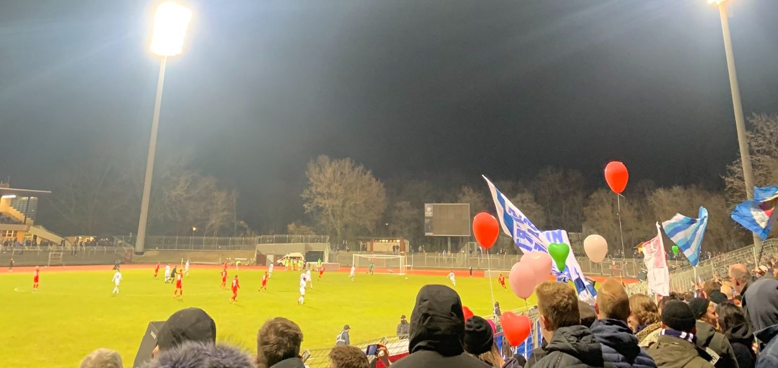 Blick in ein Stadion bei Flutlicht, im Vordergrund Fans mit Weiß-blauen Fahnen und Herzchen-Luftballons