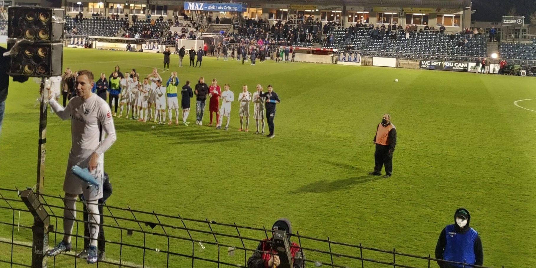 Ein Fußballspieler steht auf eiem Sicherheitszaun, im Hintergrund die Mannschaft applaudiert