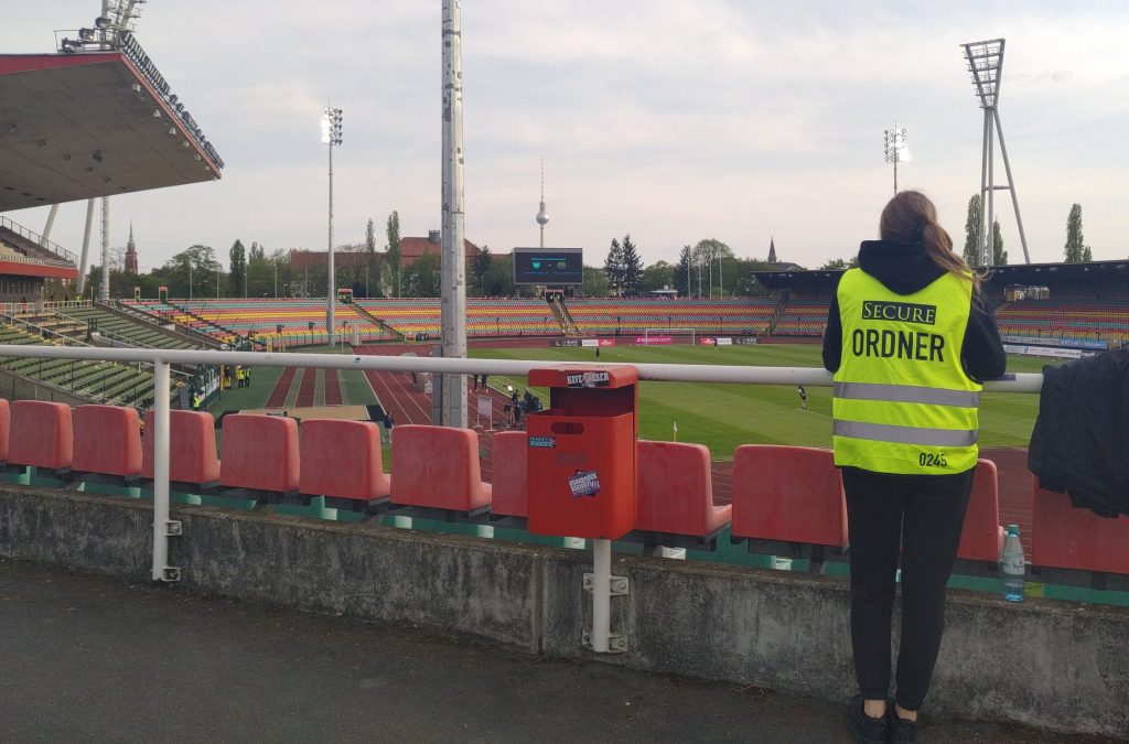 Eine Frau in Warnweste mit der Aufschrift "Ordner" schaut in ein Stadion, im Hintergrund ist der Berliner Fernsehturm zu sehen