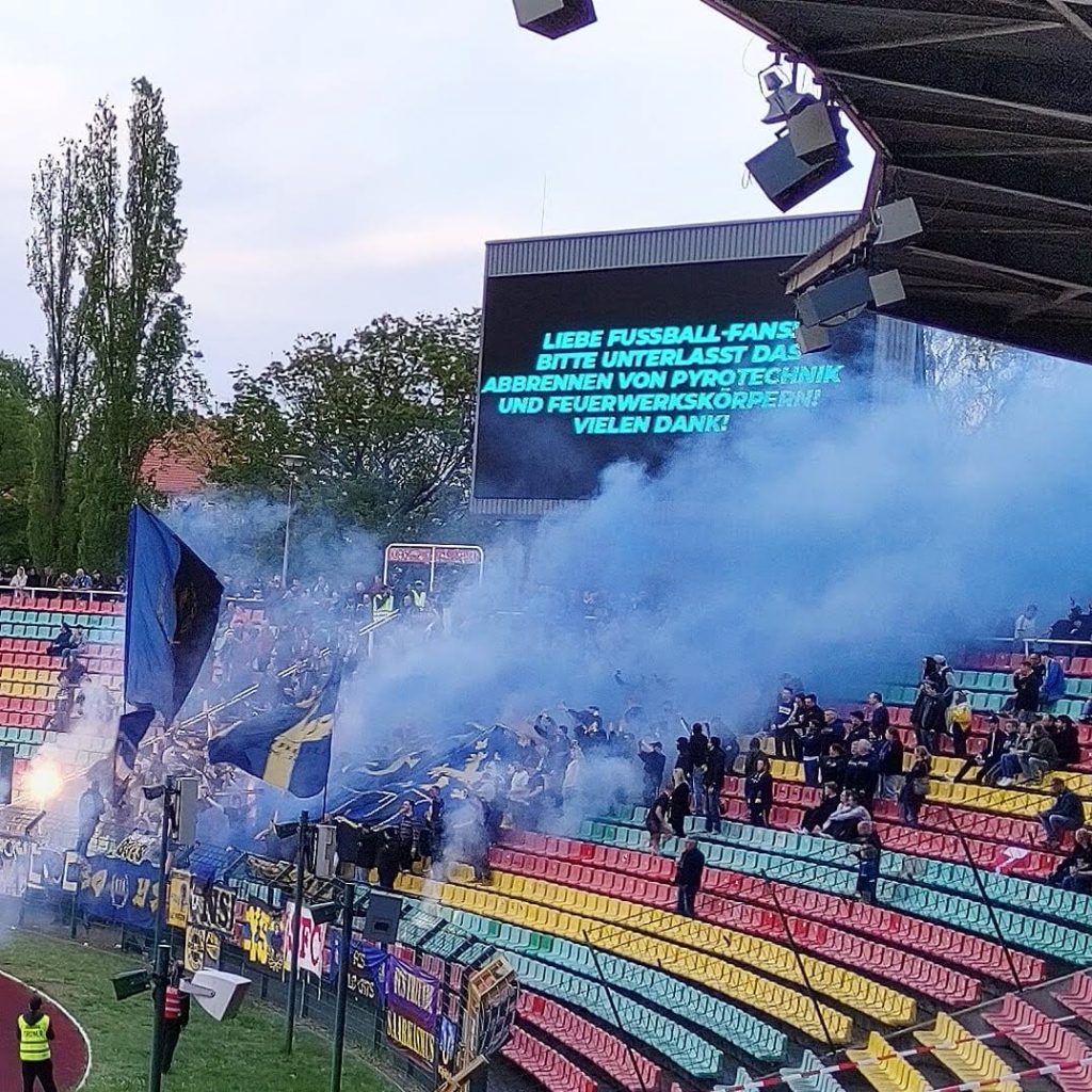 Tribüne eines Stadion. Darauf blauer Rauch, Fahnen und Fans. Im Hintergrund eine Anzeigentafel, die dazu auffordert keine Pyrotechnik abzubrennen