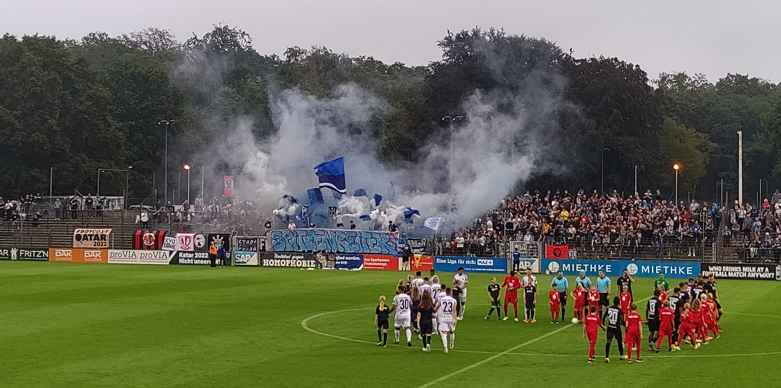 Blick in ein Fußballstadion, im Vordergrund nehmen zwei Mannschaften Aufstellung im Hintergrund weht von einer Tribüne blau-weißer Rauch herüber, dazwischen Fahnen und an der Bande ein Banner "Spitzenreiter"