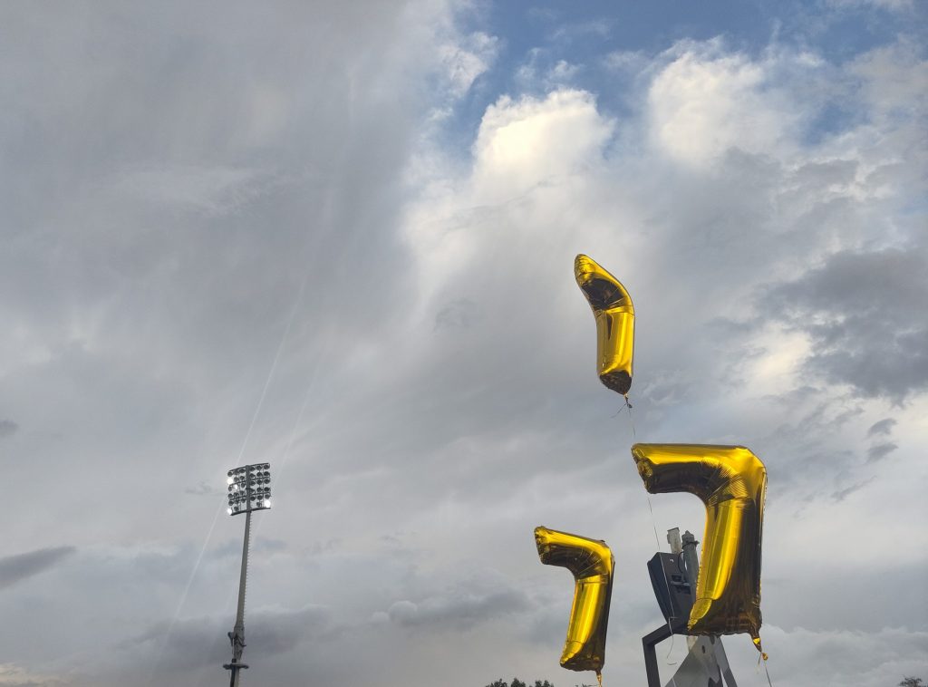 Bick in den Himmel, ein Flutlichtmast ragt hinauf, daneben drei goldfarbene Luftballons jeweils in Form einer 7