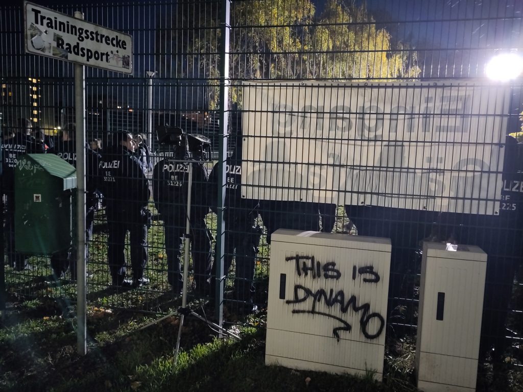 Zaun, dahinter Polizei, im Vordergrund ein Stromkasten auf dem getagged wurde "This is Dynamo"