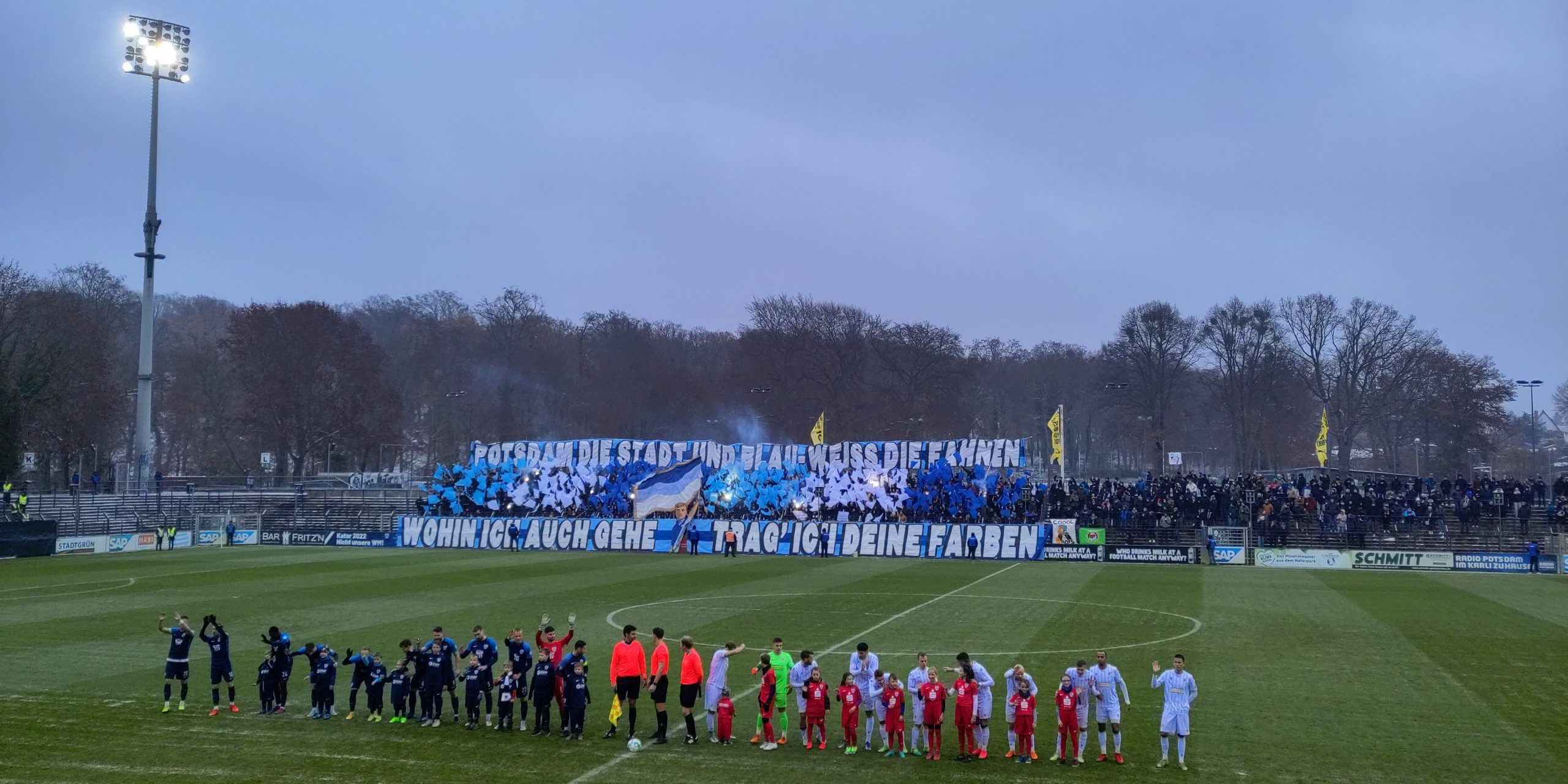 Blick auf einen Fußballplatz, im Vordergrund zwei Teams in der Aufstellung vor dem Spiel im Hintergrund aufwändige Fan-Choreo der Babelsberger Szene